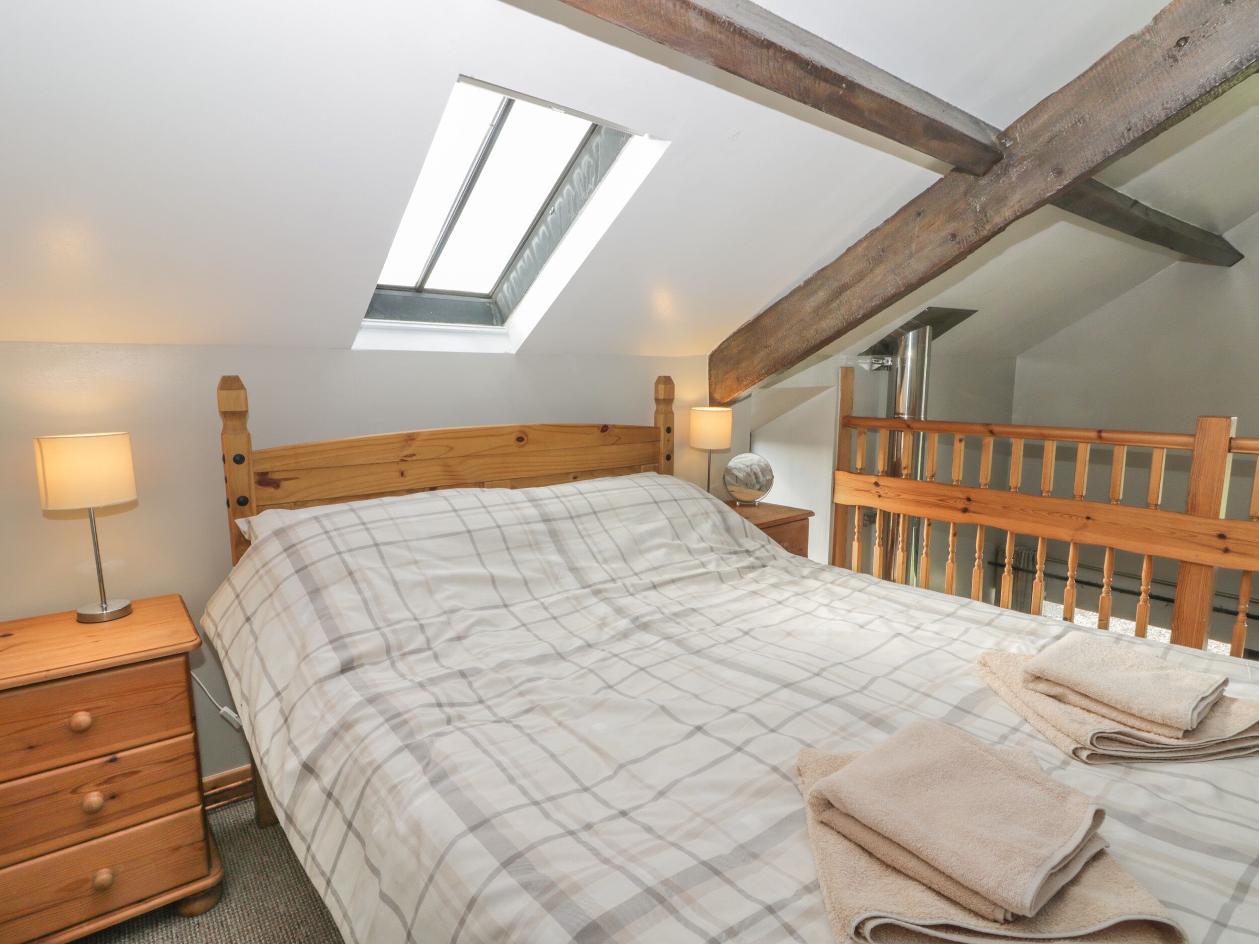 A bedroom with a sloped ceiling, a skylight, a wooden headboard, a bed with a plaid bedspread, a nightstand with a lamp, and a wooden dresser.