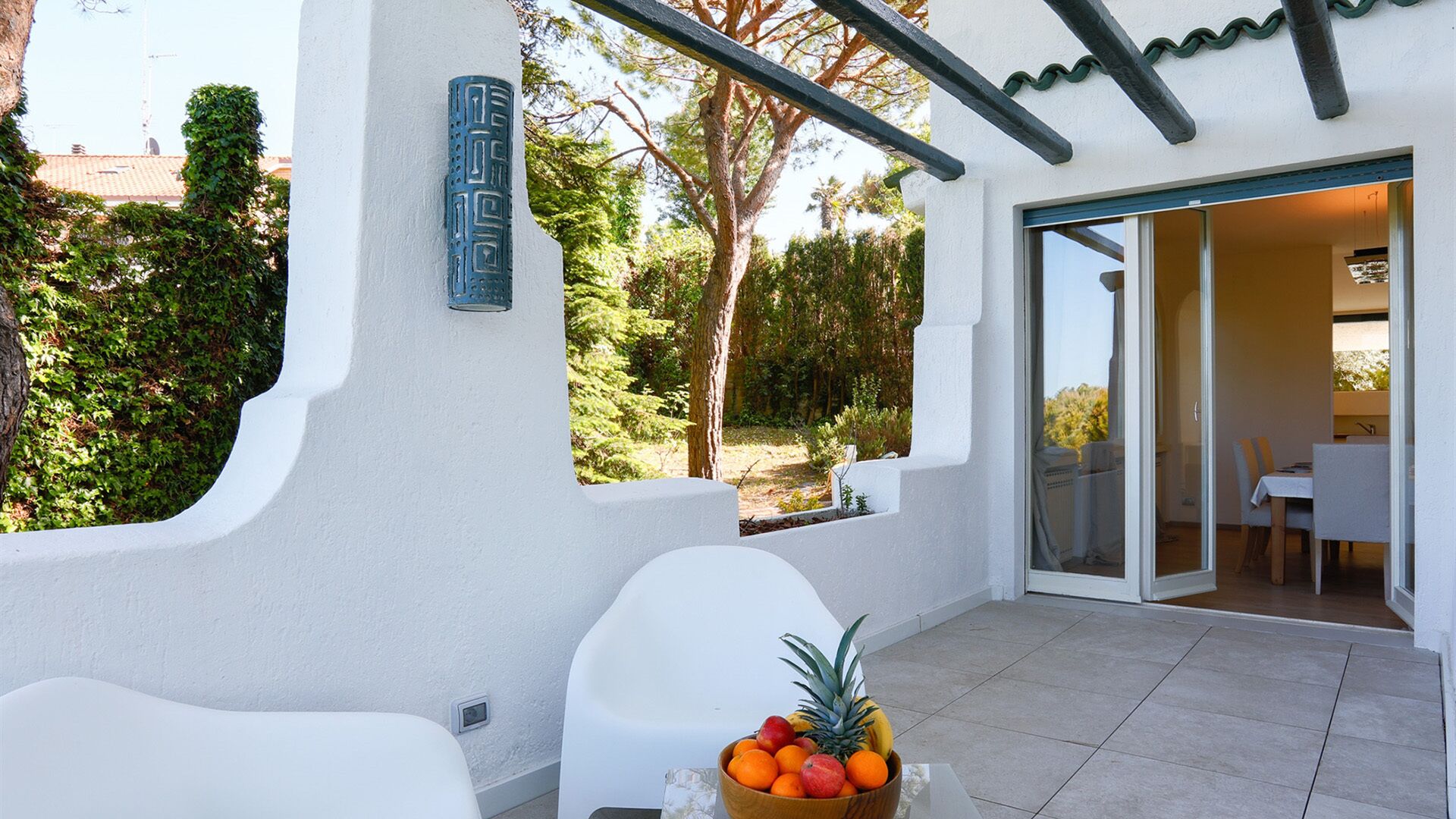 Une terrasse extérieure moderne, avec des meubles blancs, un panier de fruits et une vue sur un jardin luxuriant et l’entrée d’un immeuble.