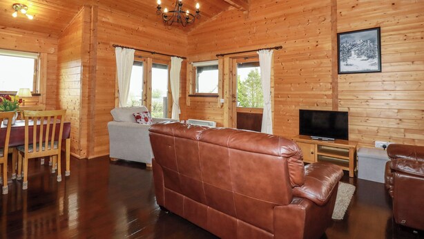 A wooden cabin interior with a leather sofa, a dining table with chairs, a TV, and a painting on the wall. - Cedar Lodge (Bodmin)