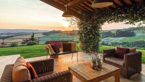 An outdoor patio with wicker furniture, a wooden table, and a view of rolling hills and a sunset.