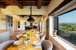 A dining area with a wooden table set for six, a view of the outdoors through a large window, and a rustic interior with exposed beams and a brick wall.