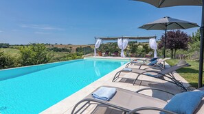 A swimming pool with lounge chairs and an umbrella, surrounded by a garden and a view of the countryside.
