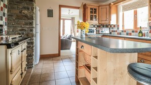 A kitchen with a stone wall, wooden cabinets, a large island, and a view to a living area. - Blaen Henllan (Builth Wells)