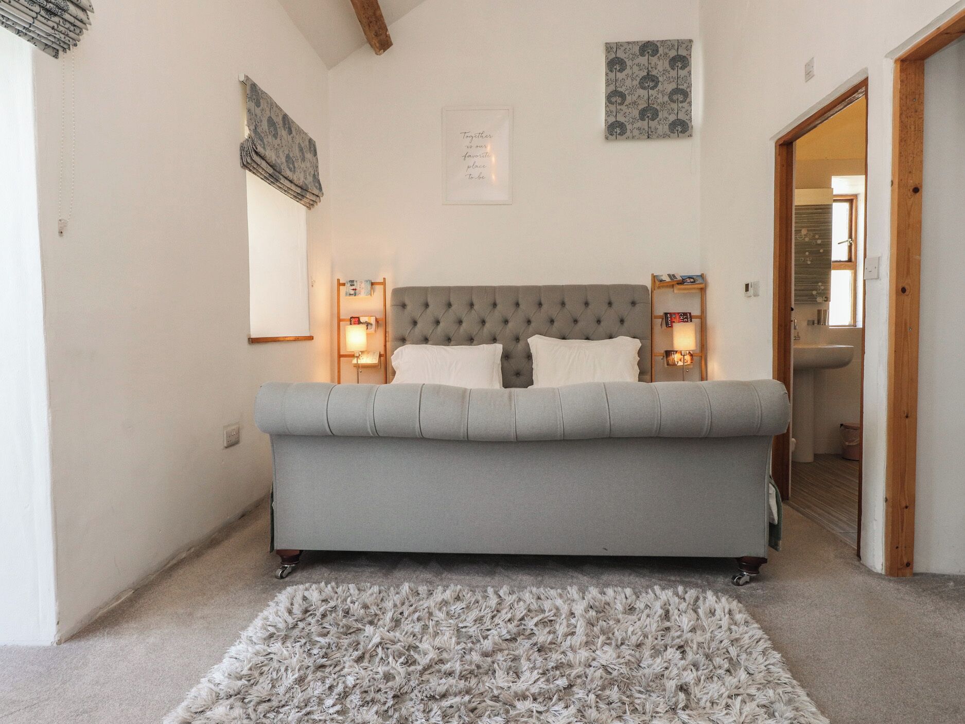 Een slaapkamer met een gestoffeerd hoofdeinde, een grijze bank, een zacht tapijt, bedlampen en een citaat in een lijst op de muur.