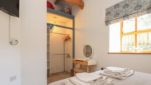 A bedroom with a bed, a desk, a mirror, a window with blinds, and a closet with hanging clothes. - Wern Olau Cottage (Caernarfon)