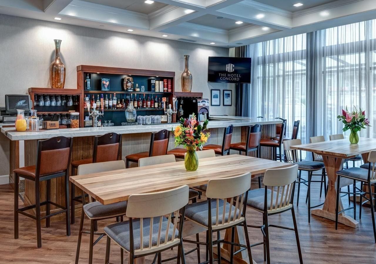 Un bar de hotel con mesas y sillas de madera, una barra con varias botellas y una pantalla grande que muestra el nombre del hotel.