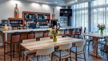 Un bar de hotel con mesas y sillas de madera, una barra con varias botellas y una pantalla grande que muestra el nombre del hotel.