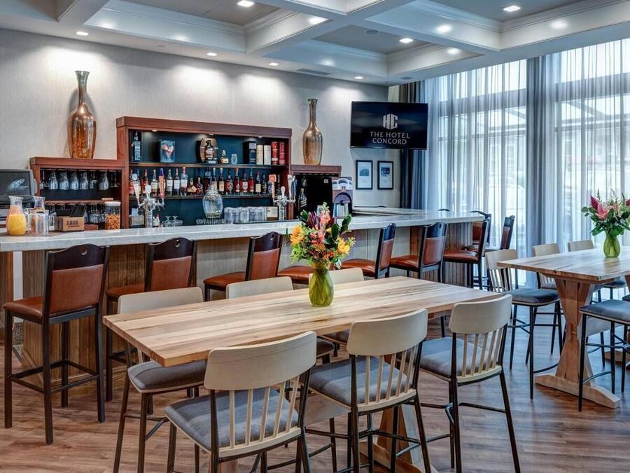 Un bar de hotel con mesas y sillas de madera, una barra con varias botellas y una pantalla grande que muestra el nombre del hotel.
