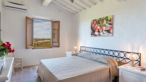 Une chambre à coucher avec un lit, une table de chevet, une commode, une fenêtre avec un volet ouvert, un tableau représentant des pommes et un vase avec des fleurs.