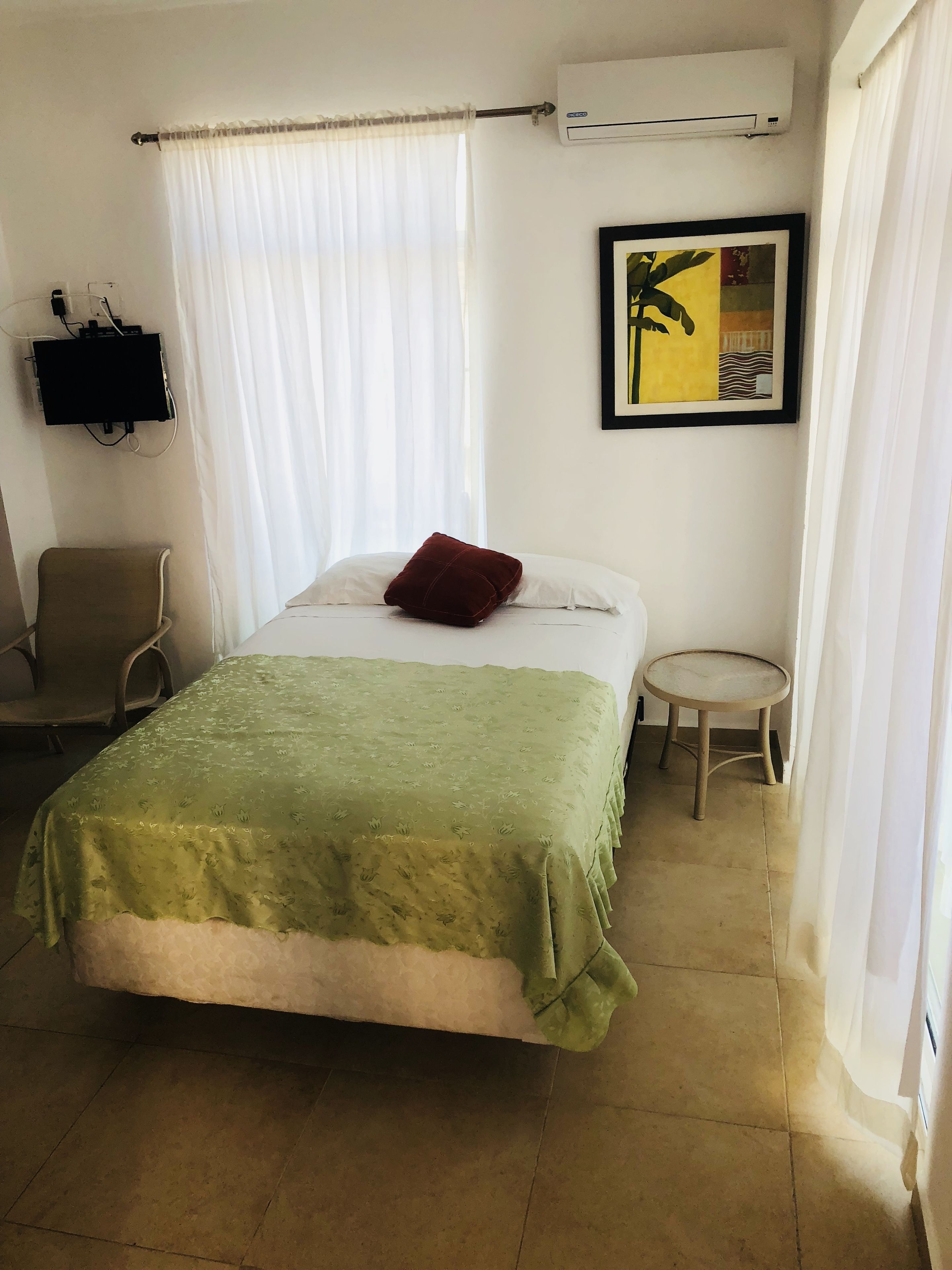 A hotel room with a bed, a chair, a small table, an air conditioning unit, and a wall-mounted TV.