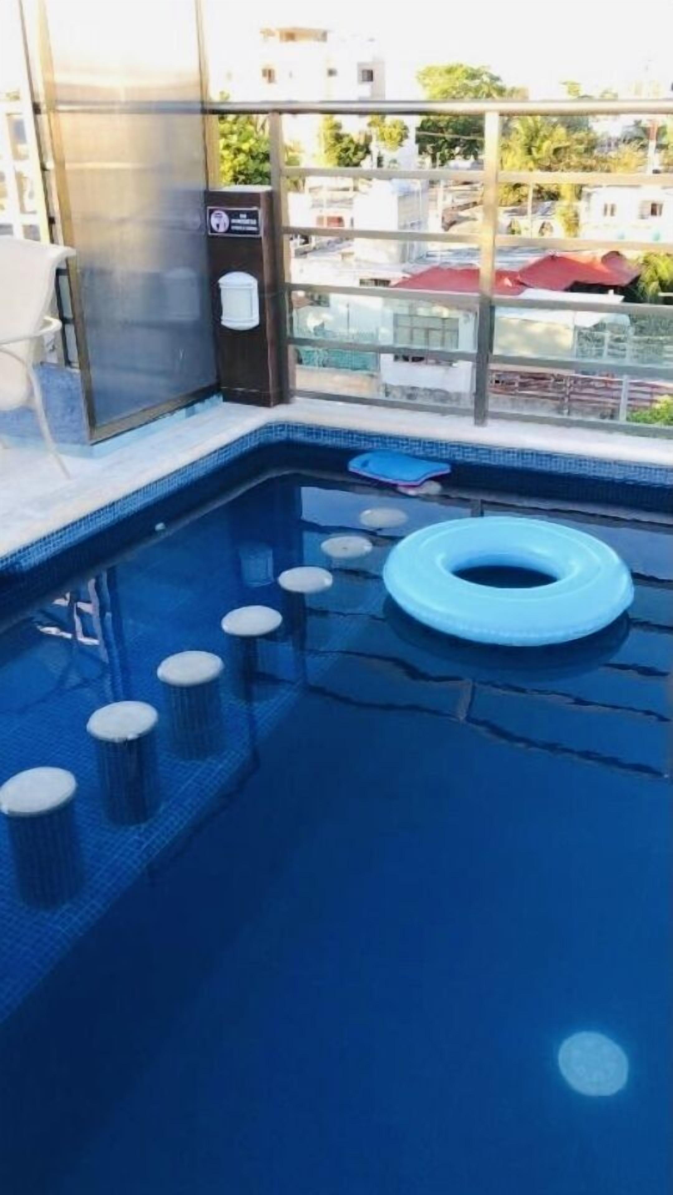 Ein Swimmingpool mit einem blauen Luftreifen, Stufen zum Wasser und Gebäuden sowie Bäumen im Hintergrund.