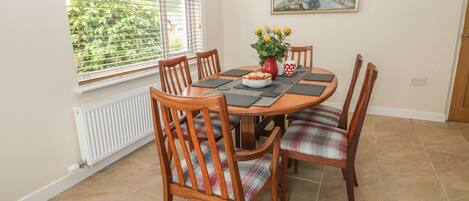 A dining area with a round wooden table, six wooden chairs with plaid cushions, a vase with yellow flowers, and a painting on the wall.