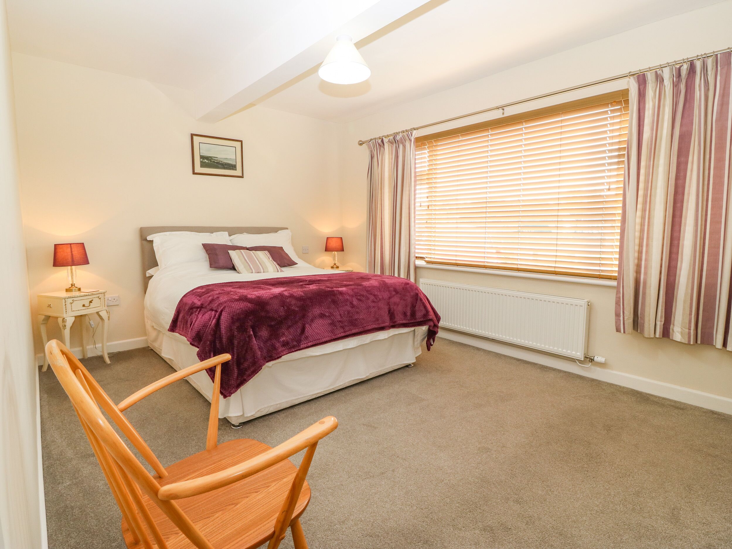 A bedroom with a bed, two bedside lamps, a window with blinds, a radiator, and a wooden chair.