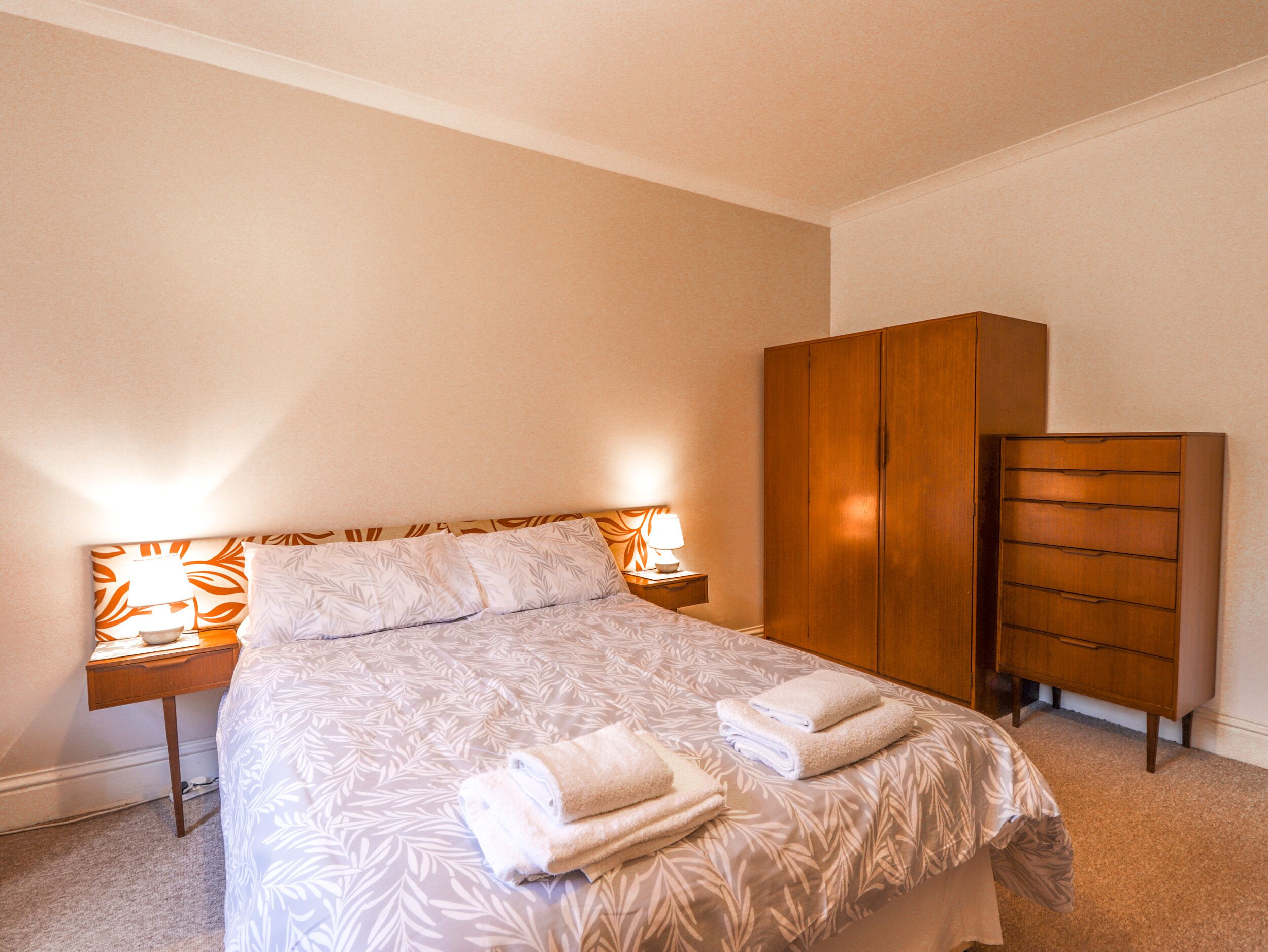 A neatly made bed with a patterned bedspread and two pillows, flanked by bedside lamps with orange lampshades. A wooden wardrobe is positioned to the right of the bed.