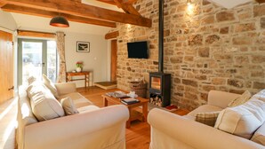 A cozy living room with stone walls, wooden beams, and a fireplace. There are two sofas and a coffee table with books and a vase of flowers. - The Byre, Sedbury Park Farm (Richmond)