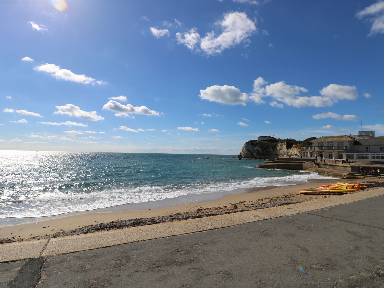 A coastal view with a sandy beach, a rocky cliff, and a building with a balcony overlooking the sea.
