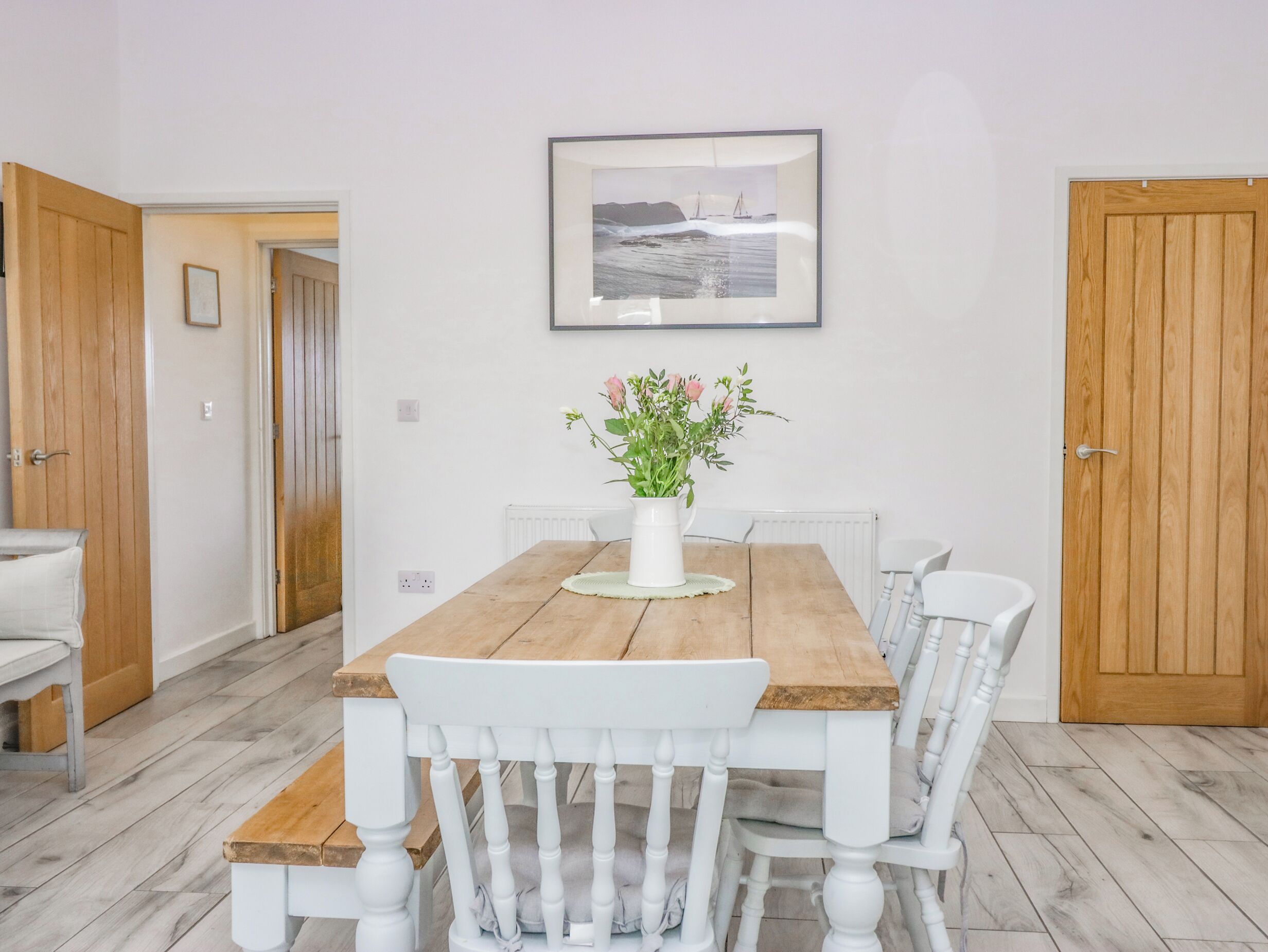 A dining area with a wooden table and white chairs, a white vase with flowers, and a framed picture on the wall.