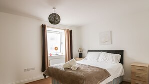 A bedroom with a bed, a nightstand, a lamp, a window with curtains, and a radiator. - 4 Bredon Court (Newquay)