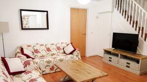 A living room with a floral sofa, a wooden coffee table, a television on a stand, and a staircase. - 4 Bredon Court (Newquay)