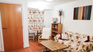 A living room with a wooden coffee table, a floral sofa, a bookshelf, and a dining area with a table and chairs. - 4 Bredon Court (Newquay)