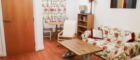A living room with a wooden coffee table, a floral sofa, a bookshelf, and a dining area with a table and chairs.