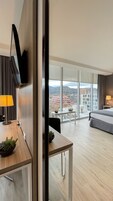Una habitación de hotel moderna con un ventanal grande, un televisor de pantalla plana montado en la pared, un escritorio de madera con una lámpara, una cama y una mesita con una planta.