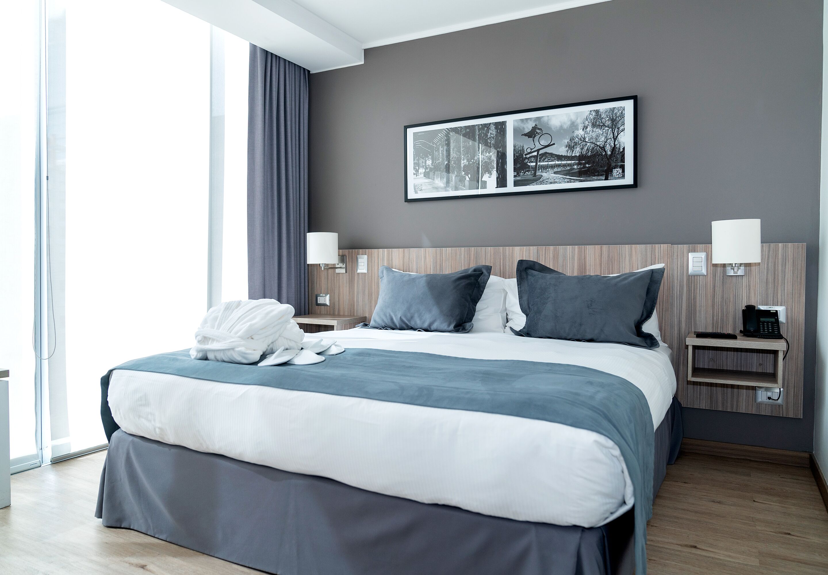 A modern hotel room with a large bed, two bedside tables, a wall-mounted telephone, and a large window with curtains.