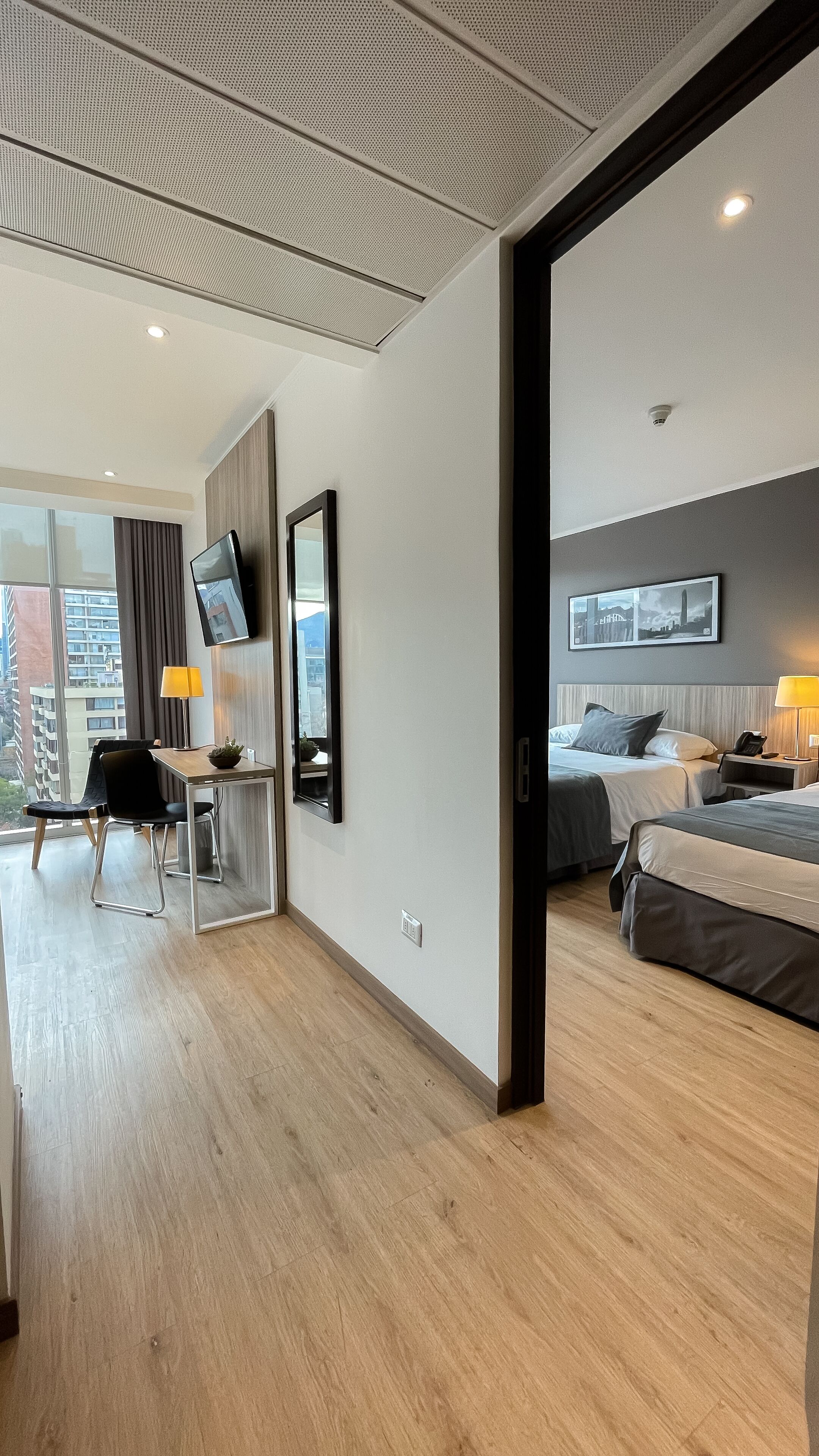 Una habitación de hotel moderna con suelo de madera, un escritorio con silla, una cama y un televisor fijado a la pared.