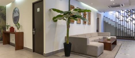 A modern lobby with a reception desk, a large potted plant, a sofa, and a staircase in the background.