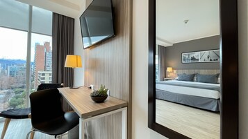 Habitación de hotel con una cama grande, un escritorio con silla, televisor montado en la pared y vistas de la ciudad a través de amplias ventanas.