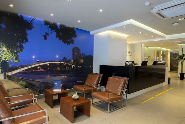 A hotel lobby with a reception desk, seating area, and a mural of a cityscape at night. - Hotel Capital Bellet (Santiago)