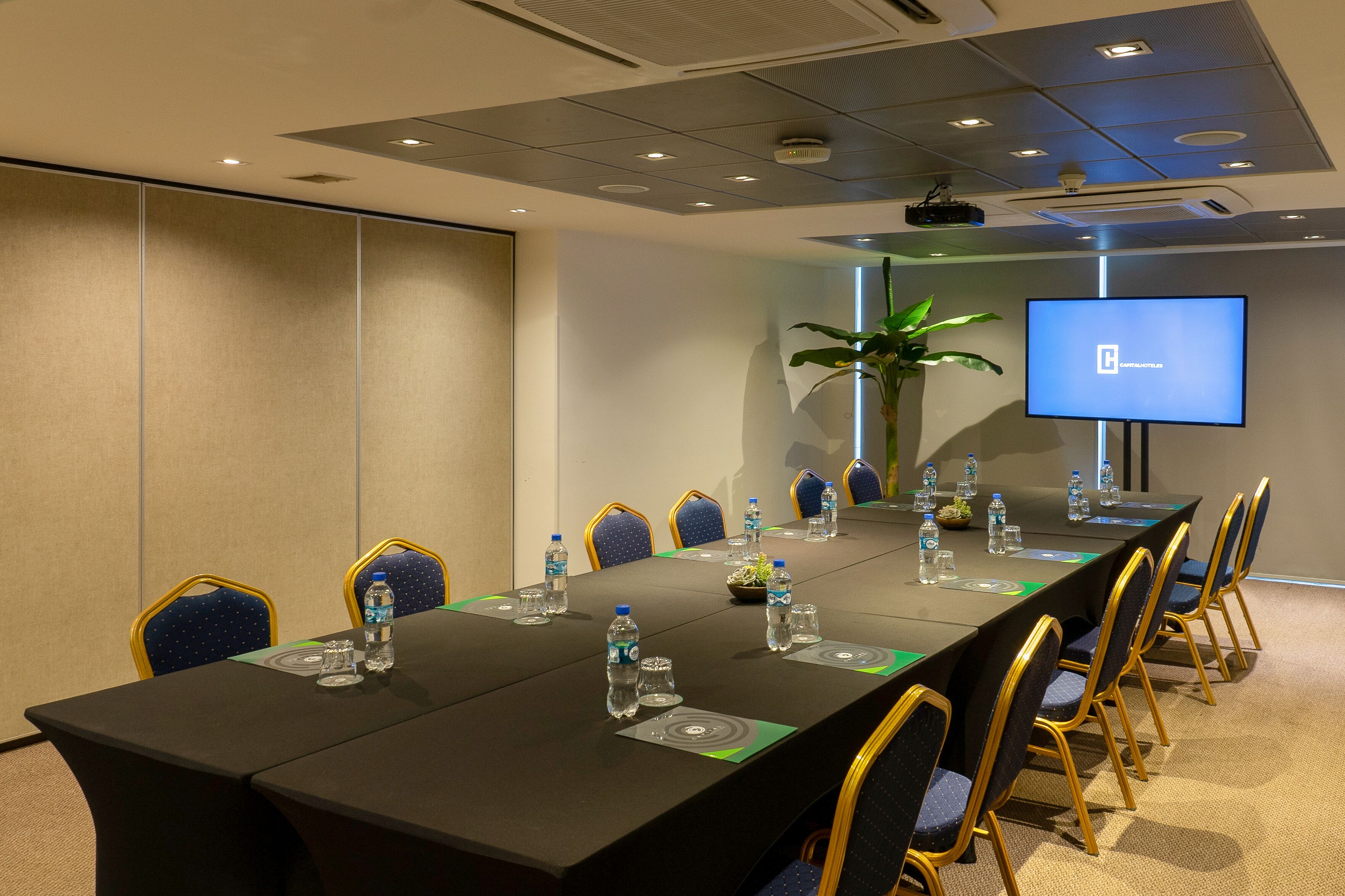 Un salón de conferencias con una larga mesa, sillas, botellas de agua y una pantalla de proyector.