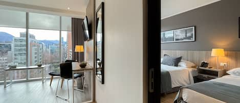 A hotel room with a large window, a desk with a chair, a bed with a bedside table, and a lamp.