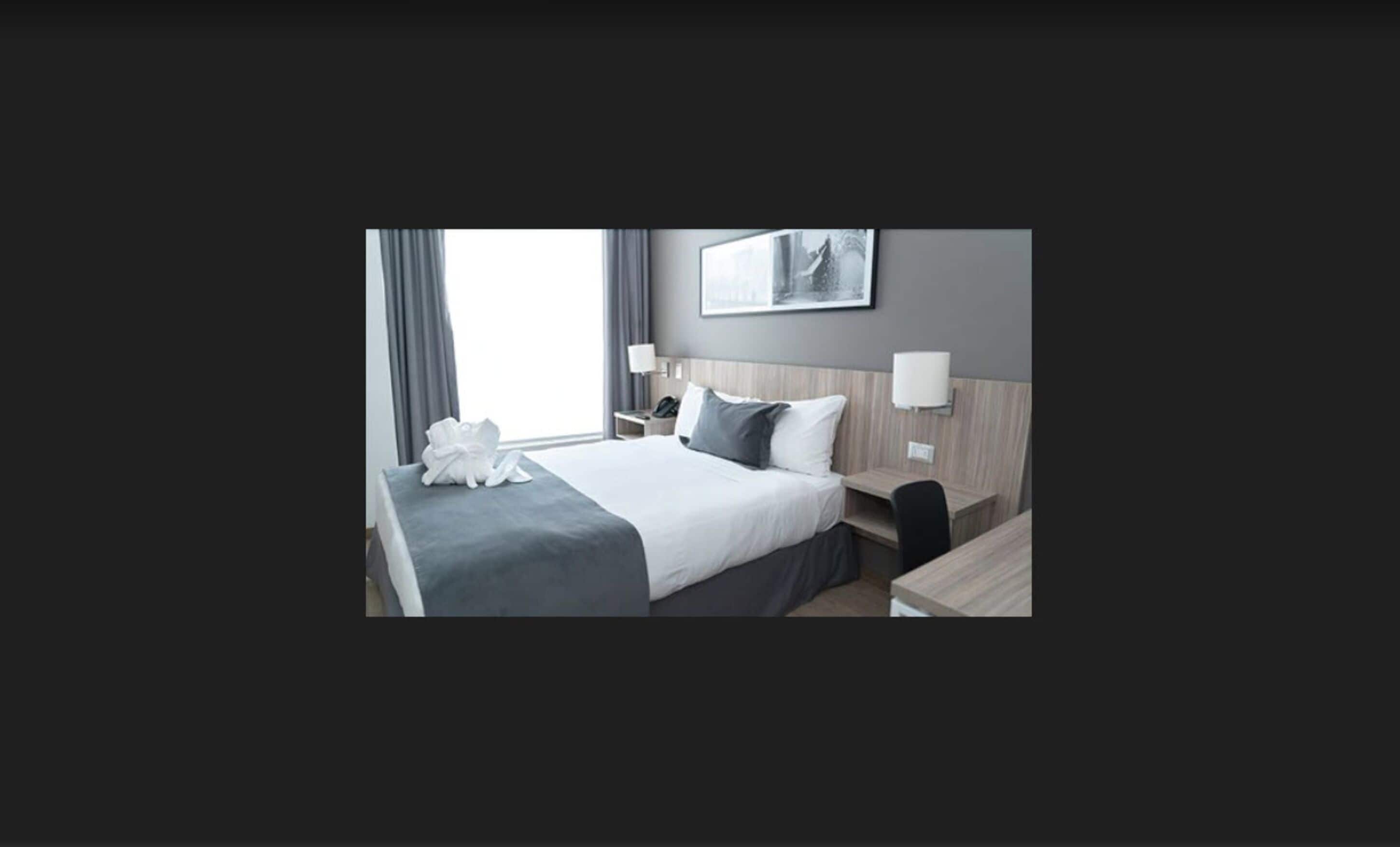 Une chambre d’hôtel moderne avec un lit bien fait, une table de chevet avec une lampe, un petit bureau avec une chaise et une grande fenêtre avec des rideaux.