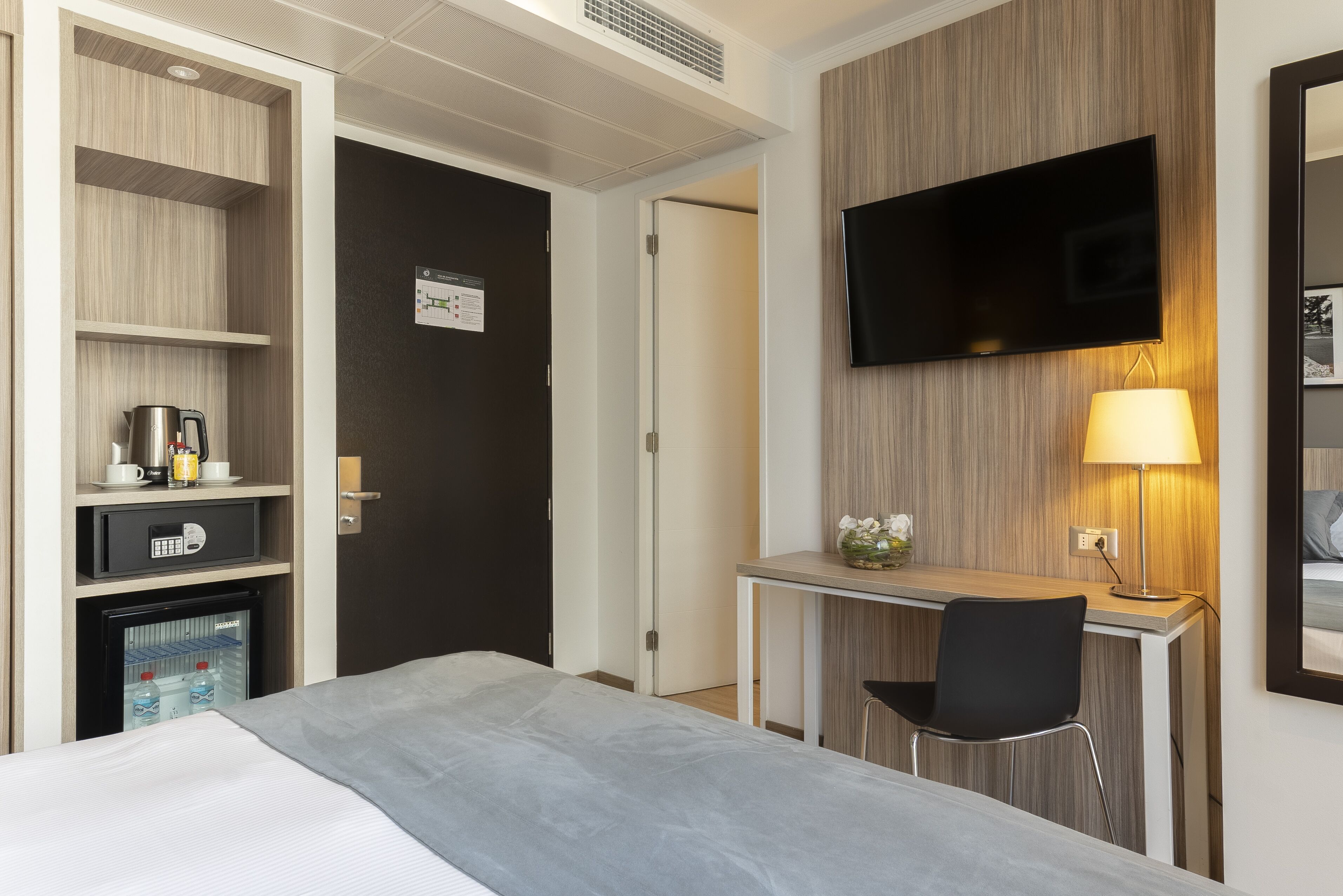 Quarto de hotel com cama, escrivaninha com cadeira, televisão, mini-geladeira e frigobar.