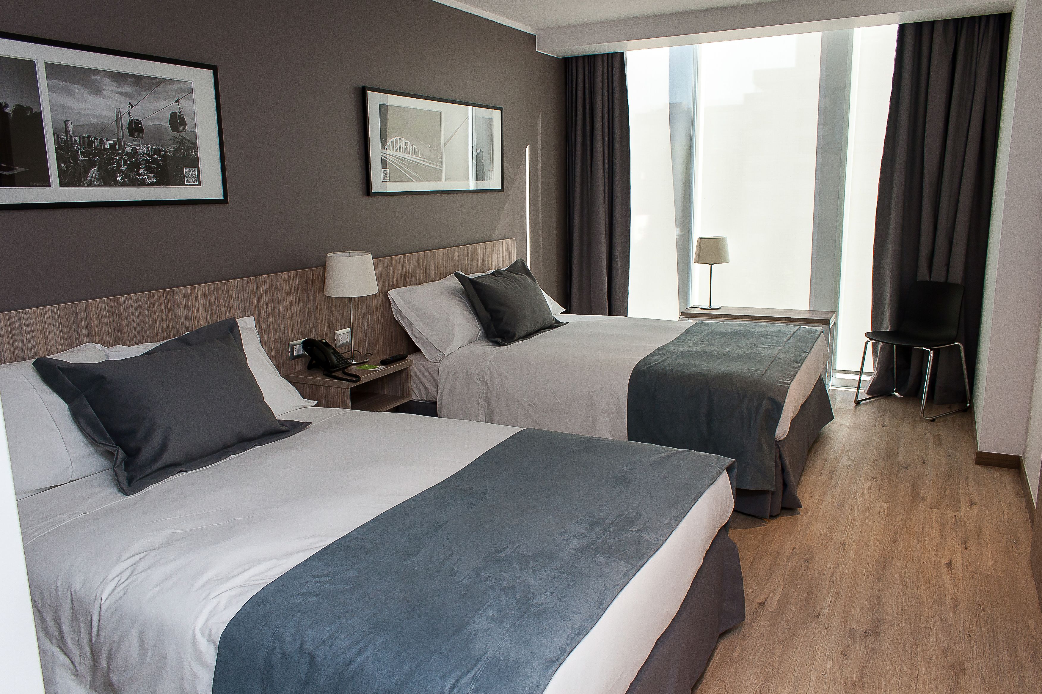 Habitación de hotel con dos camas, una mesita de noche, una silla y un ventanal con cortinas.