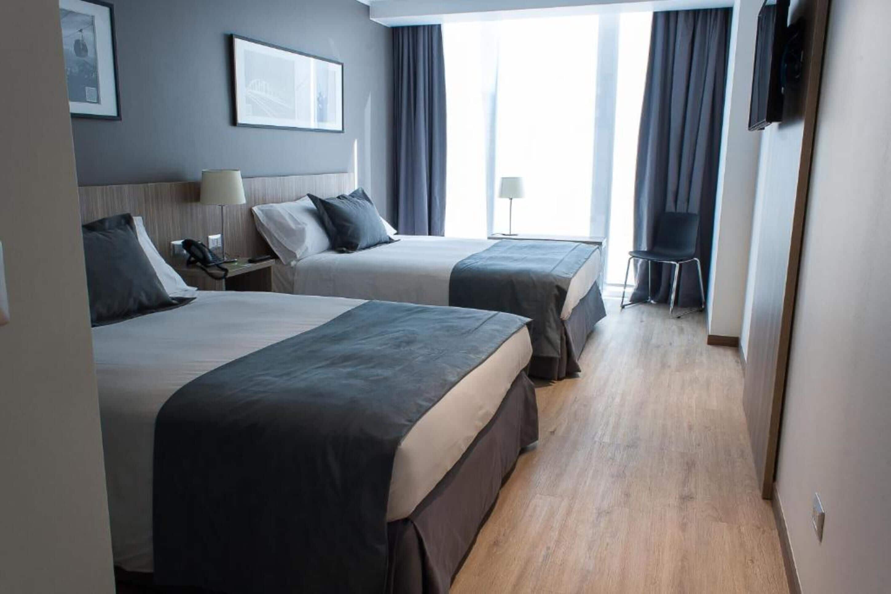 Habitación de hotel con dos camas, un escritorio con lámpara, una silla y un televisor montado en la pared.