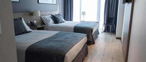 Quarto de hotel com duas camas, uma mesa de trabalho com abajur, uma cadeira e uma televisão fixada na parede.
