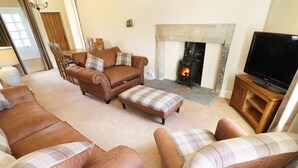 A living room with leather sofas, a plaid ottoman, a fireplace, and a television on a wooden stand. - Mill Dam Farm Cottage (Lancaster)