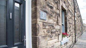 A stone building with a black door and a window with a white frame. A sign on the wall reads "3 Seashall Cottage."