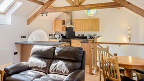 A modern kitchen with wooden beams, a dining area with a leather chair, and a view of the kitchen through a skylight. - Moor View Chapel (Camelford)