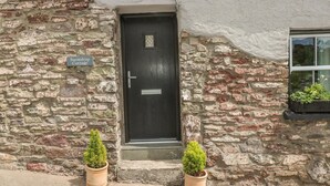 Une petite maison en pierre avec une porte sombre, une enseigne indiquant « Snowdrop Cottage », et deux plantes en pot posées sur le sol.