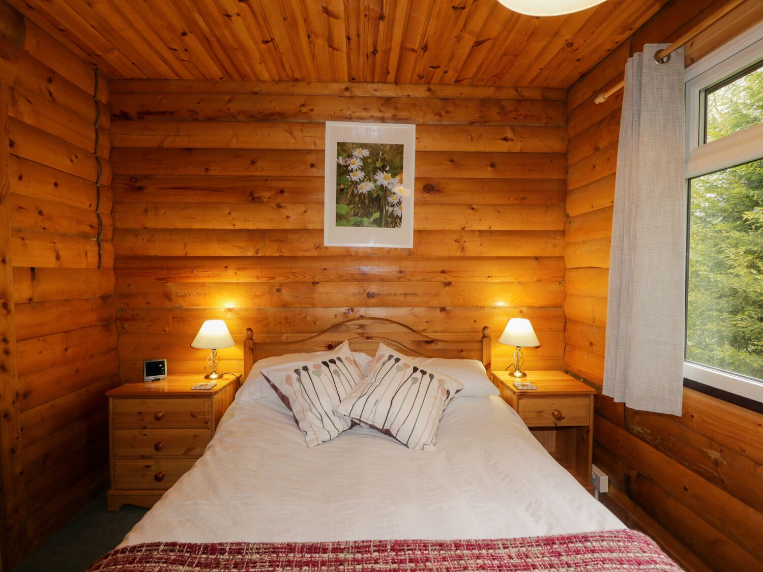 A wooden cabin bedroom with a bed, two bedside lamps, a nightstand, and a window with curtains.