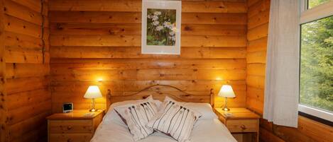 A wooden cabin bedroom with a bed, two bedside lamps, a nightstand, and a window with curtains.