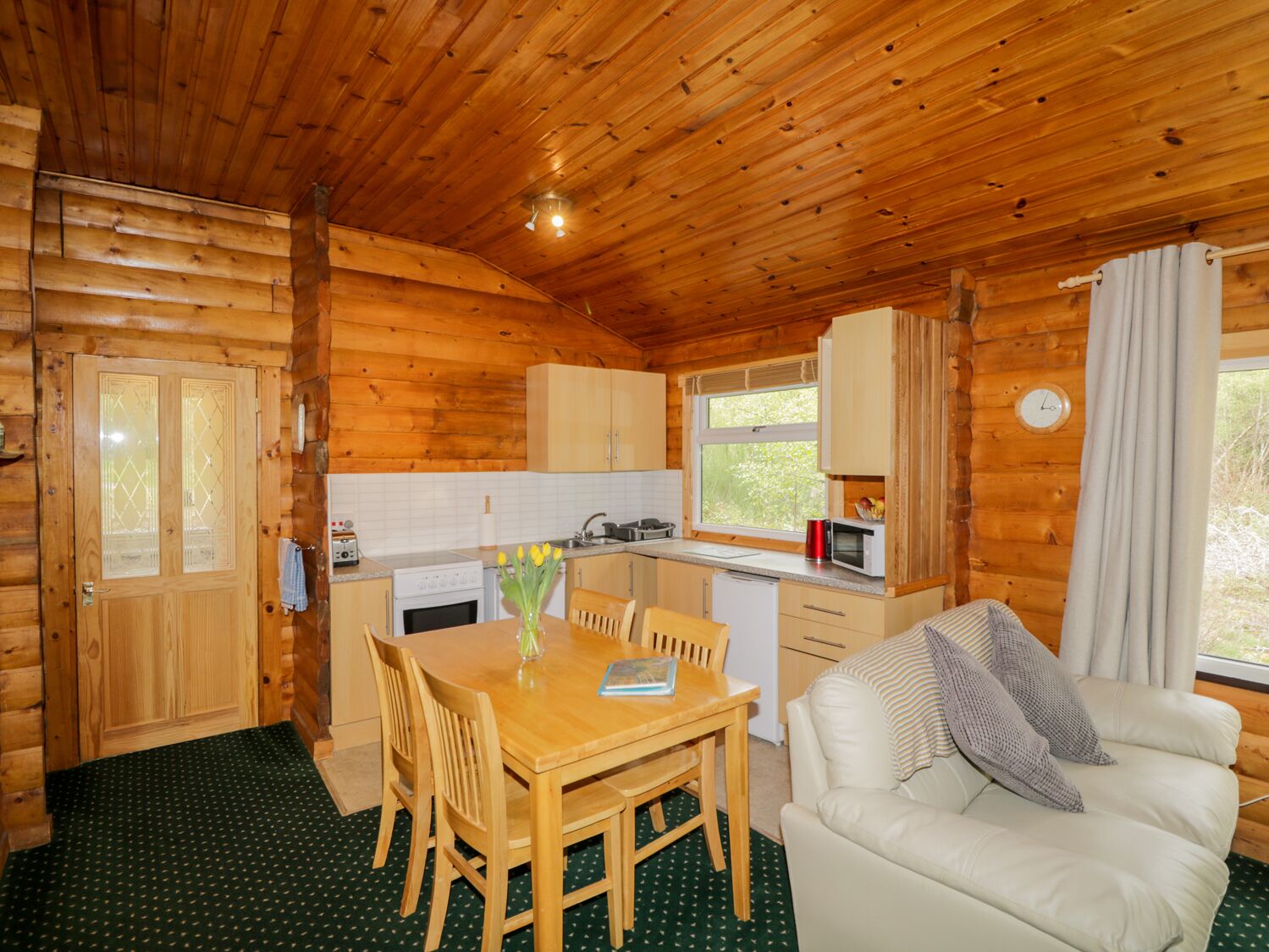 Interior acogedor de una cabaña con una mesa de comedor de madera, sillas, un sofá blanco, una zona de cocina con microondas y una ventana con cortinas.