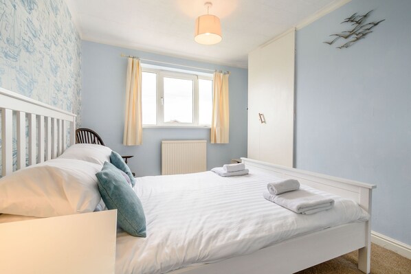 A bedroom with a white bed, a window with curtains, a radiator, and a wall-mounted light fixture.