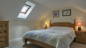 Un dormitorio con techo inclinado, tragaluz, cama de madera con colcha, dos mesitas de noche con lámparas y un aparador de madera.