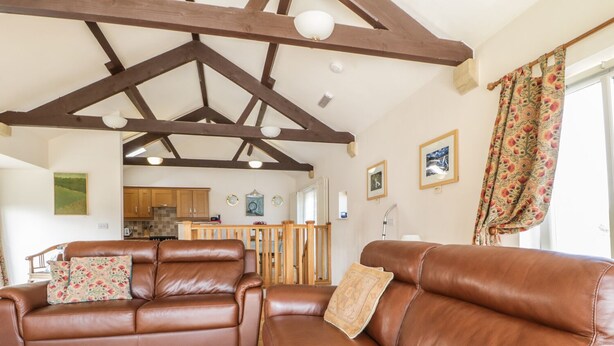 A spacious living room with two brown leather sofas, wooden flooring, and a high ceiling with exposed beams.
