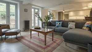 En moderne stue med en sofa, et sofabord og en lænestol. Der er en radiator på væggen og et vindue med udsigt til grønne omgivelser.