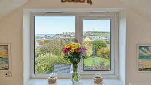 Ein Zimmer mit großem Fenster mit Blick auf eine Küstenstadt, einer Vase mit Blumen und einer Kordeldekoration an der Wand.